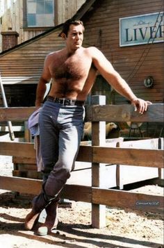 a shirtless man leaning on a fence in front of a barn with his hand on the rail