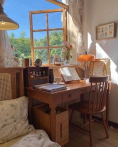 a wooden desk sitting under a window next to a chair and table with a book on it