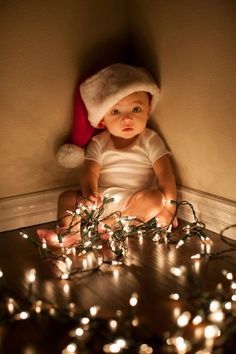 a baby in a santa hat sitting on the floor with christmas lights all around him