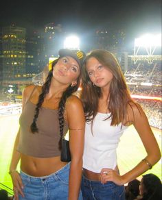 two women standing next to each other in front of a baseball field with lights on