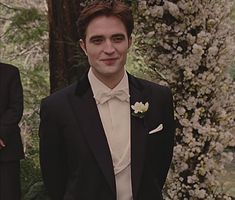 two men in tuxedos standing next to each other with trees and flowers behind them