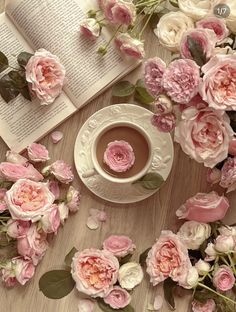 a cup of coffee surrounded by pink flowers and an open book on a wooden table