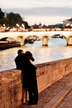 two people leaning against a stone wall next to the water