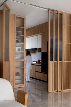 a person standing in a kitchen next to a white couch and wooden cupboards with glass doors