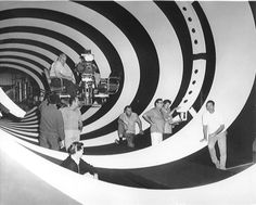 black and white photograph of people walking in an art gallery with large spiral design on the wall