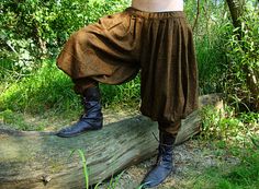 a man in brown pants and black boots standing on a tree trunk with his hands behind his head