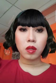 a woman with short black hair and red shirt is taking a selfie in front of an orange wall