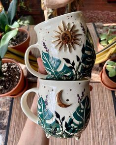 two coffee mugs that have been decorated with flowers and leaves on them, sitting next to each other in front of potted plants