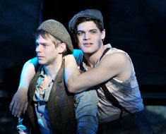 two young men sitting next to each other in front of a dark background, one with his arm around the other's shoulder