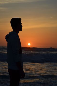 a man standing on top of a beach next to the ocean at sun setting in the sky