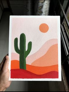 a person holding up a painting with a cactus in the desert on it's side