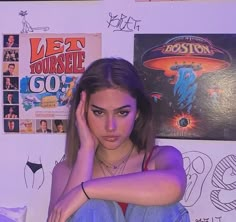 a young woman sitting in front of a wall with posters on the wall behind her