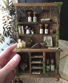 someone is holding up a miniature shelf with herbs and candles on it in front of some books