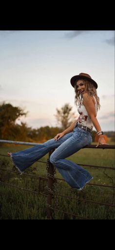 a woman sitting on top of a fence next to a green field and wearing a hat