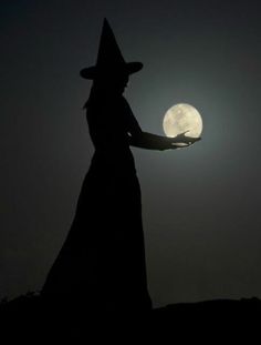 a woman in a witches hat holding the moon with her hand, silhouetted against a dark night sky