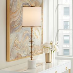 a table with a lamp, vase and flowers on it in front of a painting