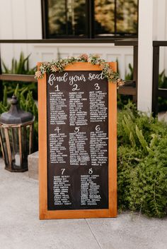 a wedding seating plan on a chalkboard in front of a building with greenery