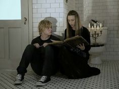 a woman sitting on the floor reading a book next to a man in a bathroom