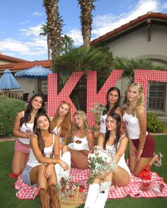 a group of women sitting on top of a blanket in front of a house with the letter k