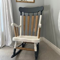 a white rocking chair sitting in front of a painting on the wall next to a fan