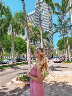 a woman in a pink dress is standing on the sidewalk near palm trees and buildings