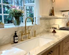 a white kitchen with marble counter tops and gold faucets, windows, and sink