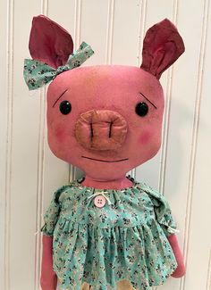 a pink stuffed pig wearing a green floral dress and bow on its head, hanging from a white wall