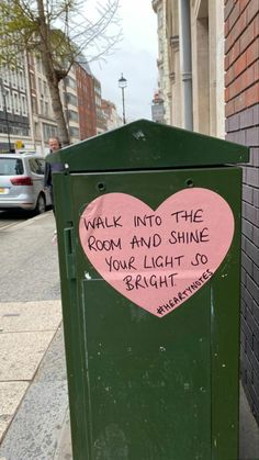 a green trash can with a pink heart on it's side next to a brick wall