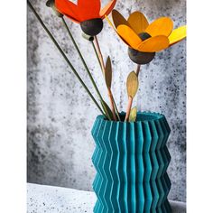 three colorful flowers in a blue vase on a white counter top with grey wall behind it