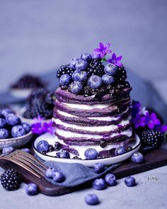 a cake with blueberries and purple flowers on top is sitting on a plate next to some berries