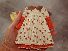 a hand holding a small doll in a white and orange dress with apples on it