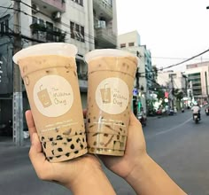 two cups of coffee are held up in the air by someone's hand on a city street