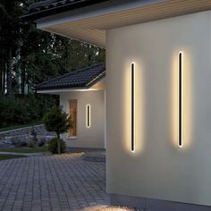 two lights are on the side of a white wall near a brick walkway and trees