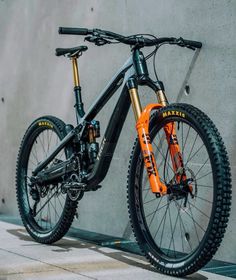 an orange and black mountain bike leaning against a concrete wall with no wheels on it