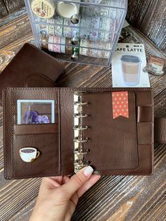 a hand holding an open brown leather planner book next to a cup of coffee and other office supplies