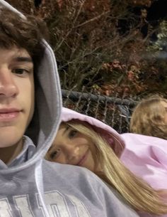 a young man and woman wearing hoodies in front of a fence with trees behind them