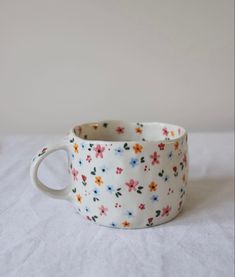 a white cup with colorful flowers on it sitting on top of a bed next to a wall