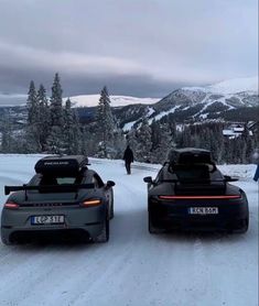 two sports cars parked on the side of a snow covered road with trees in the background