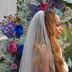 a woman with long hair wearing a wedding veil and standing in front of colorful flowers