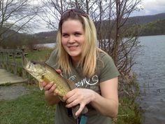 a woman is holding a fish in her hands and smiling at the camera while standing near a body of water