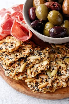 an assortment of olives and crackers on a wooden platter with a bowl of olives