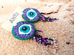 two purple and green beaded earrings laying on sand