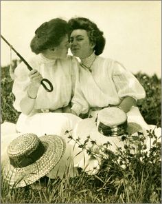 two women are sitting in the grass and one is holding a fishing pole while the other holds a hat