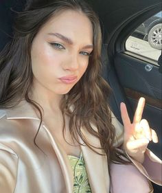 a woman sitting in a car making the peace sign with her hand and wearing a gold jacket