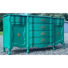 a green dresser with gold handles and drawers on it's sides, sitting in gravel