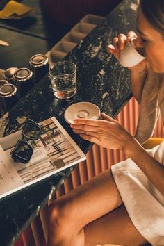 a woman sitting at a table drinking from a cup and saucer while reading a magazine