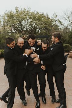 a group of men in tuxedos hugging each other on a brick walkway with trees in the background