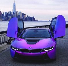 a purple car with its doors open next to the water in front of a city skyline