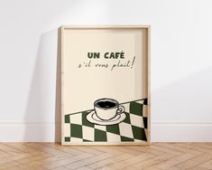 a poster with a cup of coffee on it in front of a white wall and wooden floor