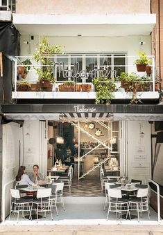 two people sitting at tables in front of a building with plants on the balconies